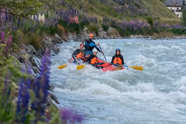 Group of rafters paddling through scenic Engadin river, with vibrant flowers lining the banks and exciting rapids ahead.