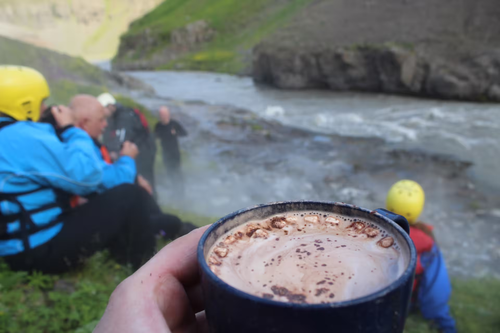 Family enjoying a river rafting break with hot chocolate, surrounded by scenic landscapes and adventure gear on a riverbank.