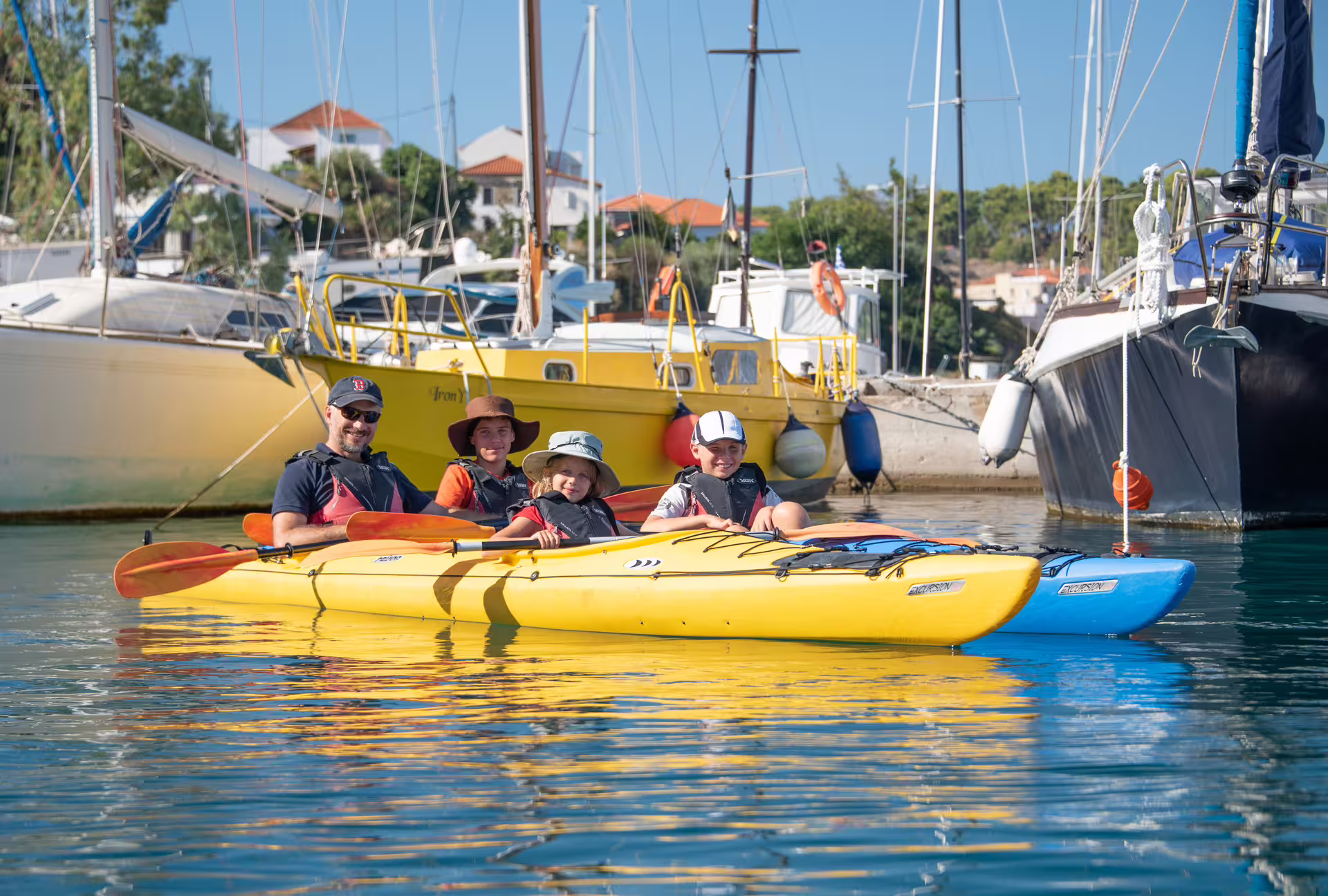Family sea kayaking in Navarino Bay marina, Pylos, with stable tandem kayaks and sailboats behind