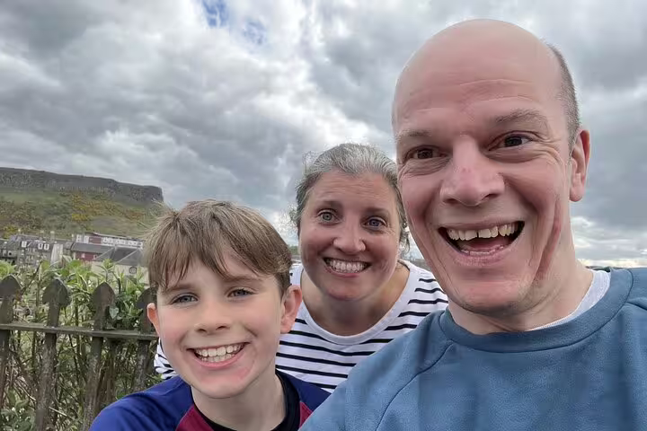 Family of three smiling on Edinburgh walking tour with rocky hillside in background