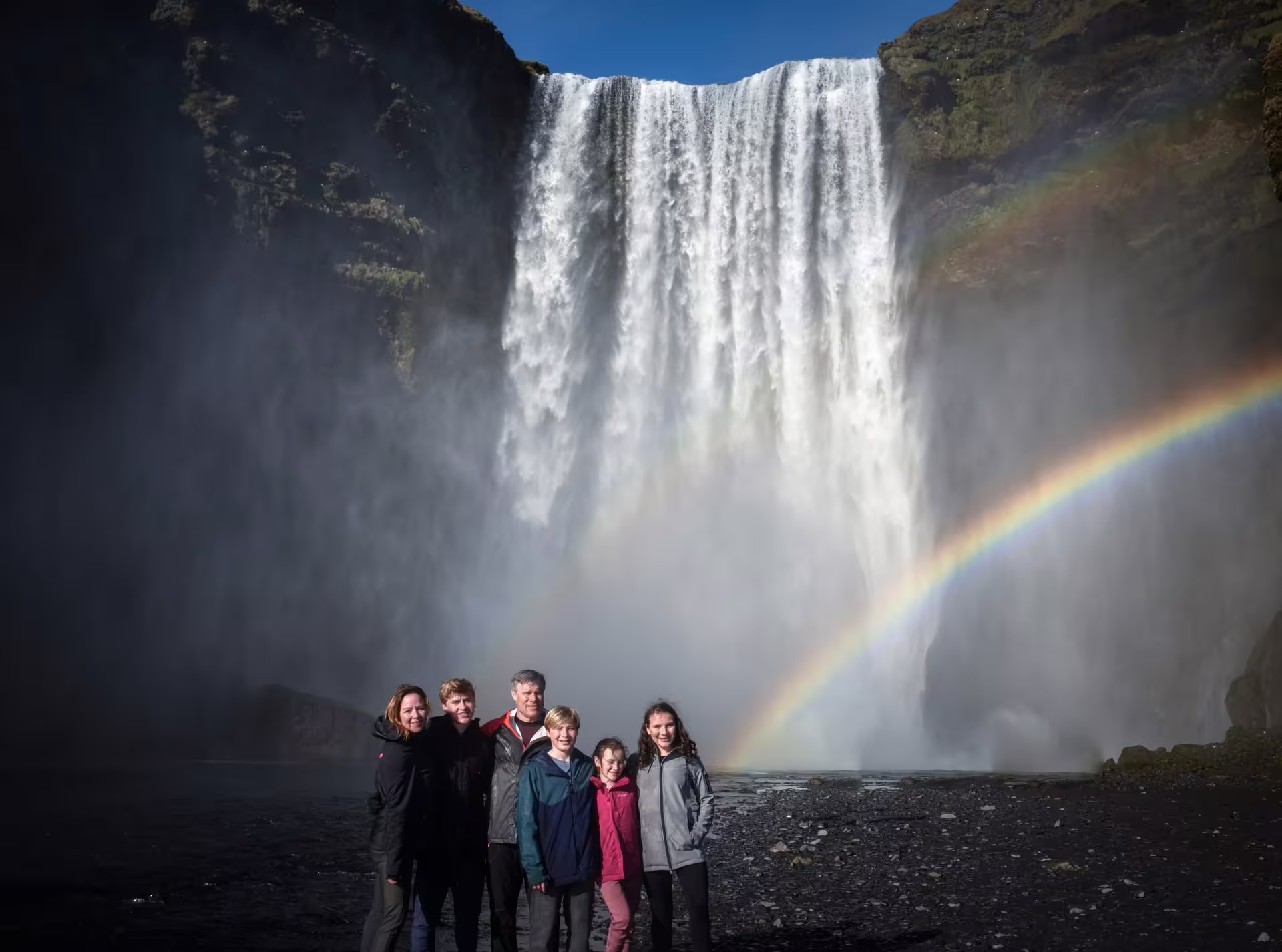 Family posing in front of Skogafoss waterfall with a vivid rainbow on Iceland South Coast captured private tour