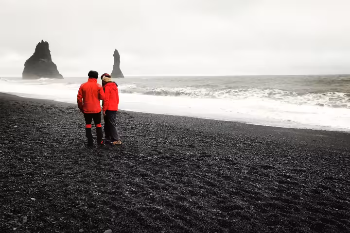Couple in red jackets standing on Iceland's black sand beach with dramatic sea stacks in background