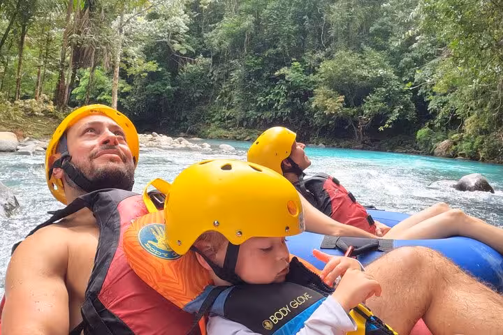 Family with helmets and life vests enjoying a serene tubing adventure on the Celeste River, enveloped by tropical greenery.