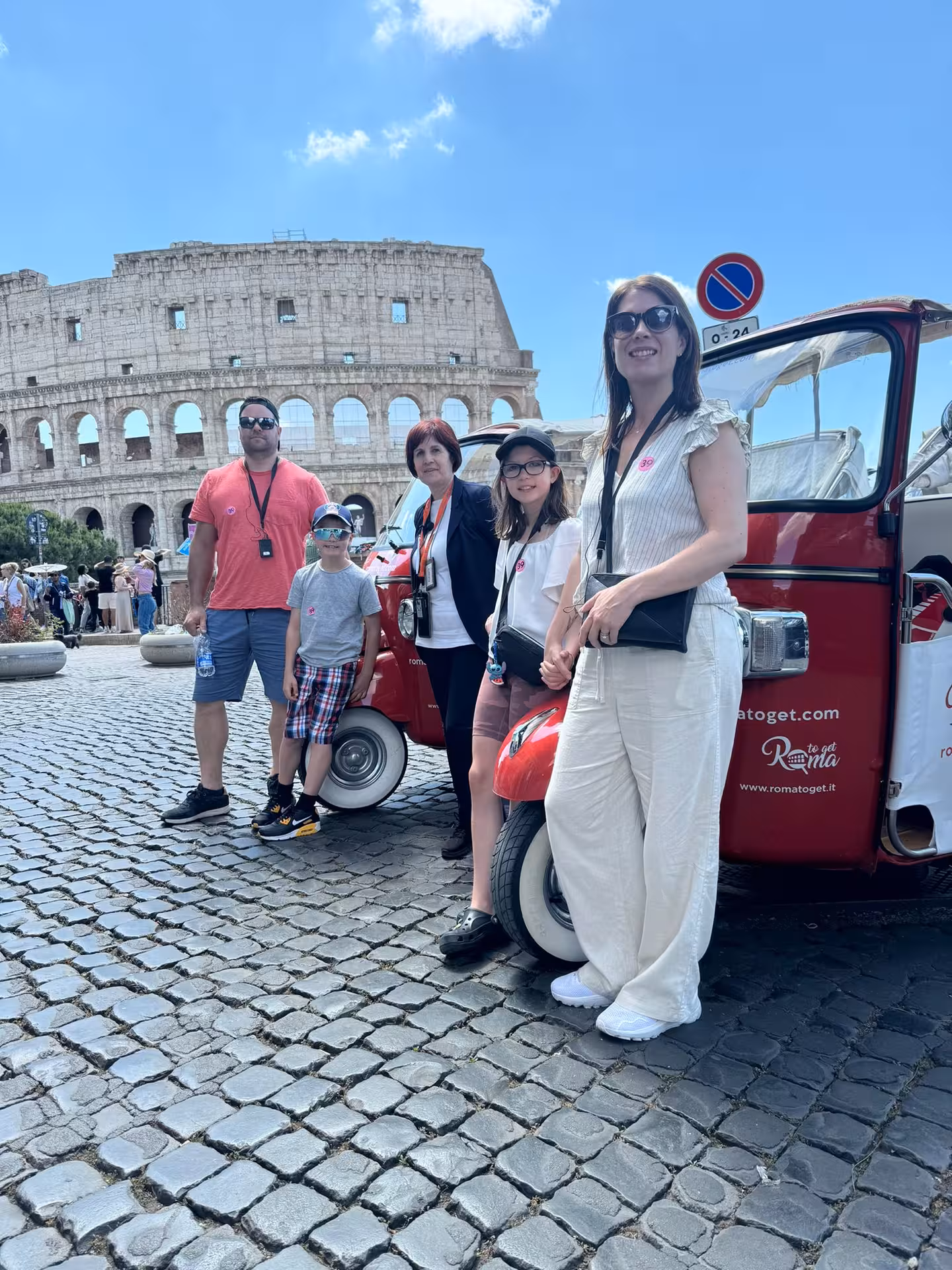 Family poses with a tuk-tuk in front of the Colosseum, experiencing a unique Rome city tour under blue skies.