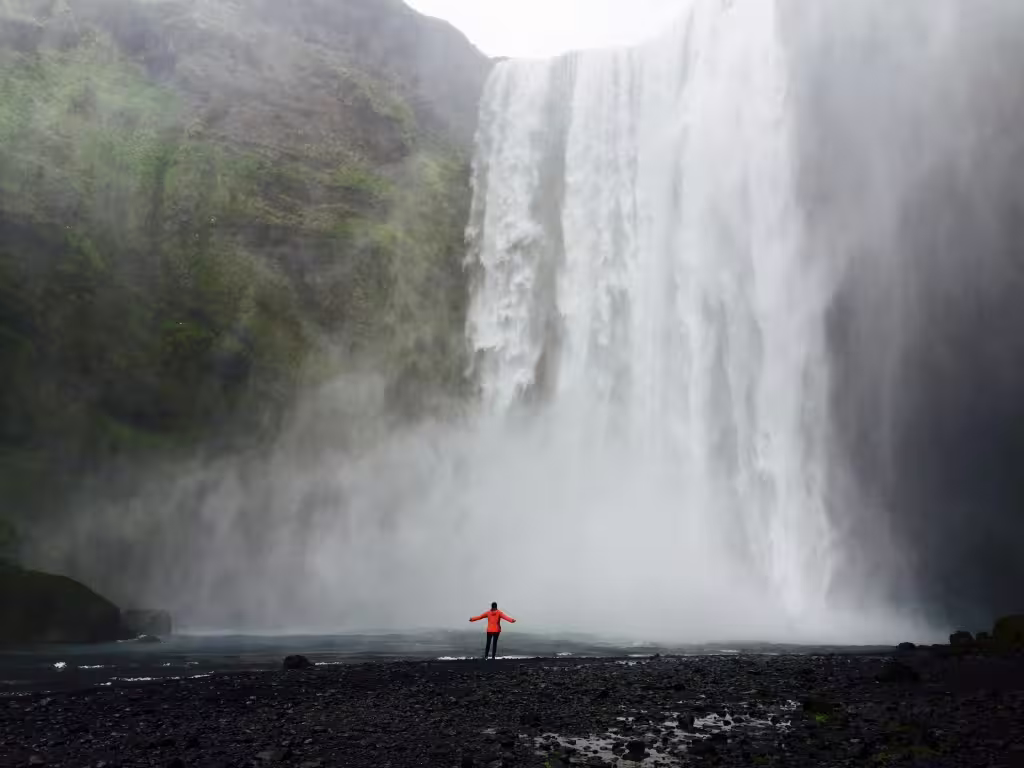 Person in red jacket standing before massive Skógafoss waterfall in Iceland surrounded by mist