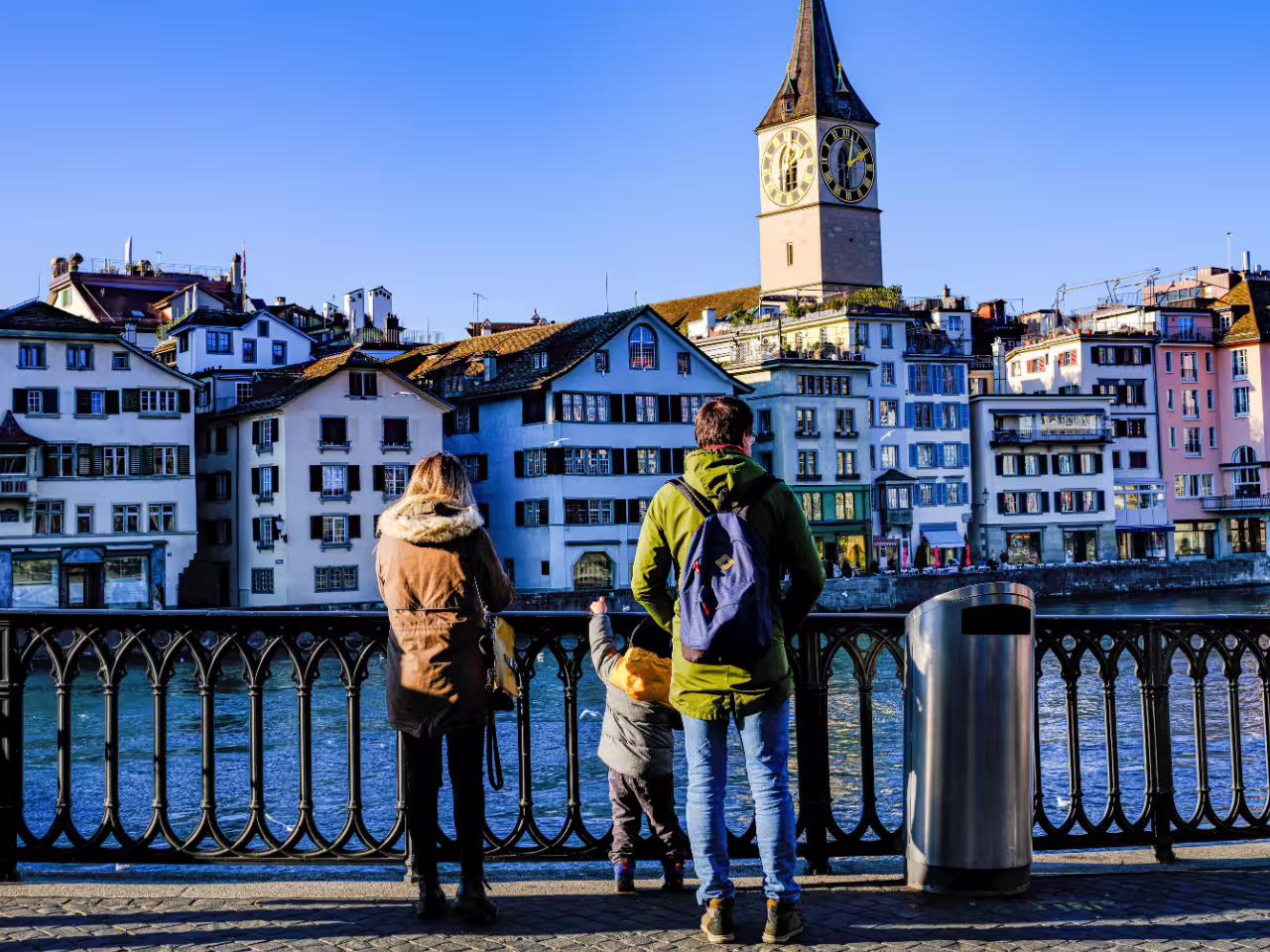 Family enjoying a scenic view of Zurich's historic buildings and clock tower by the river on a sunny day.