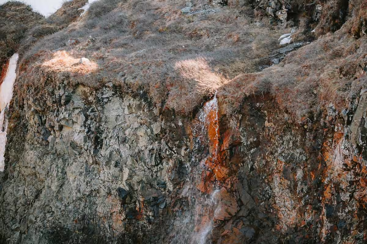 Small waterfall streaming down rugged rocky cliff face with dry grass tufts in Iceland