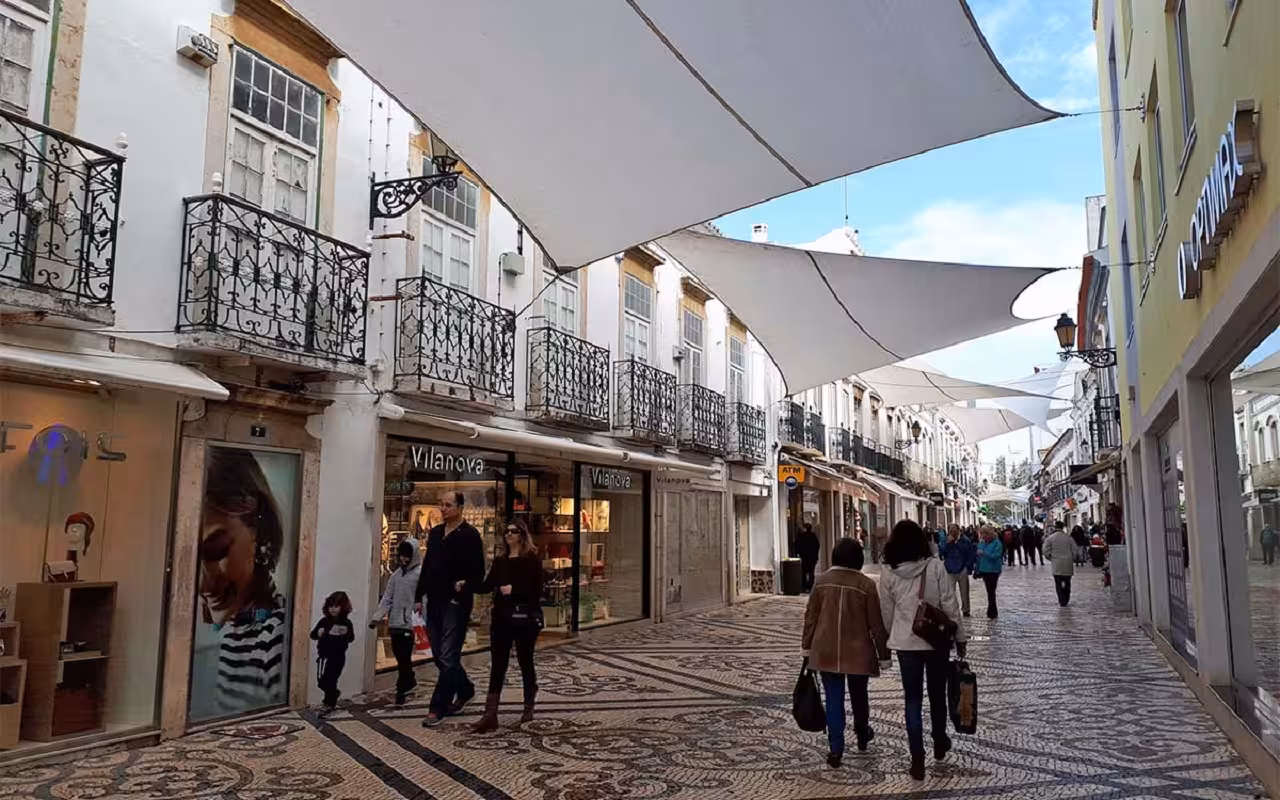 Shoppers stroll along a mosaic-paved pedestrian street in Faro old town, Eastern Algarve, shaded by white canvas sails
