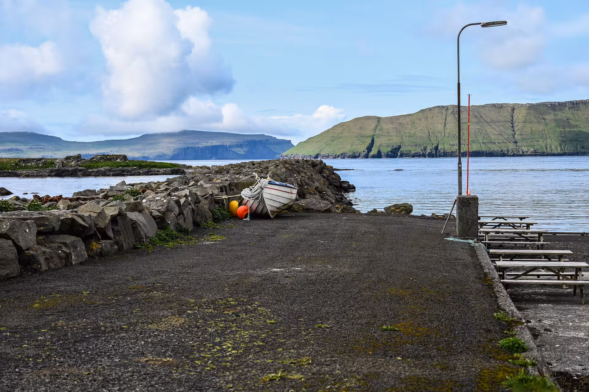 Rocky harbor pier with small white boat and colorful buoys, green cliffs and calm sea in Faroe Islands