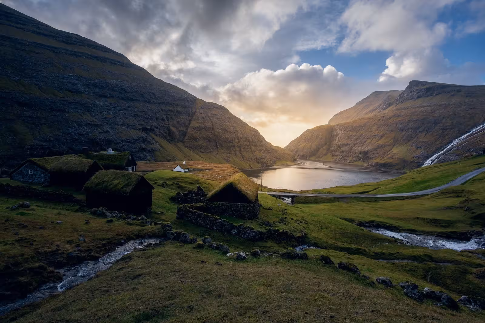 Turf-roofed village houses in Saksun valley with a lagoon and dramatic mountains at sunset, Faroe Islands