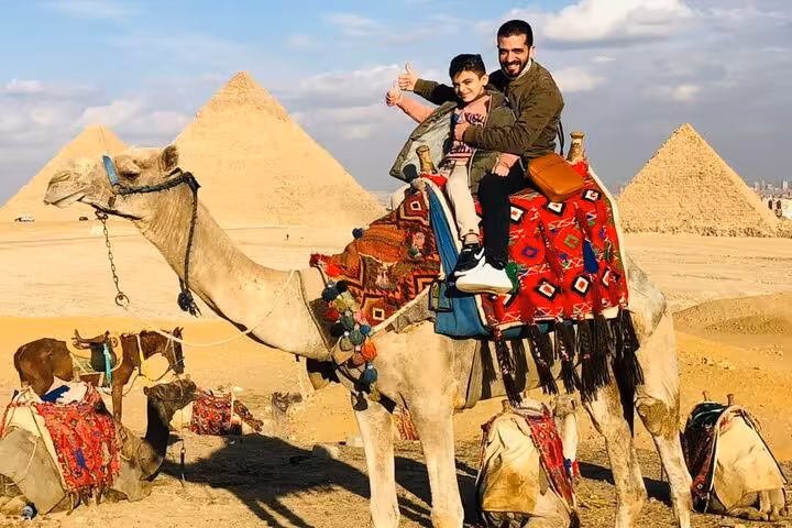 Father and son enjoy a camel ride with the Giza Pyramids behind, included in all-inclusive Giza tour package