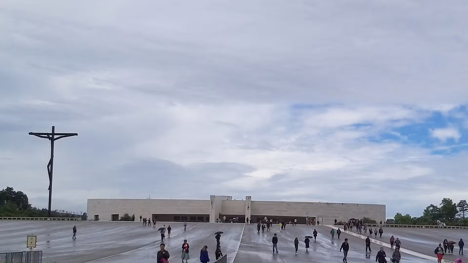 Visitors explore the expansive courtyard of Fátima Sanctuary, featuring a prominent cross under a cloudy sky.