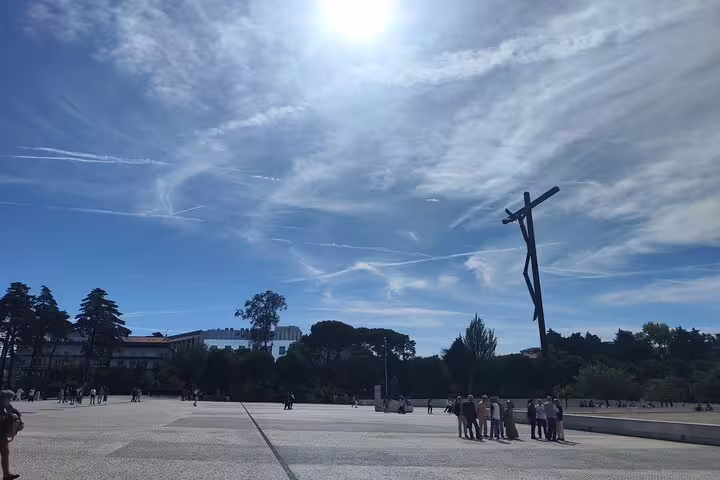 Pilgrims gather under a bright sky at Fátima Sanctuary, with a modern cross and lush surroundings, symbolizing faith and devotion.