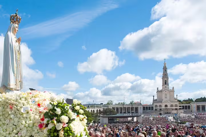 A large crowd at Fatima Shrine with the Virgin Mary statue and basilica under a bright blue sky.
