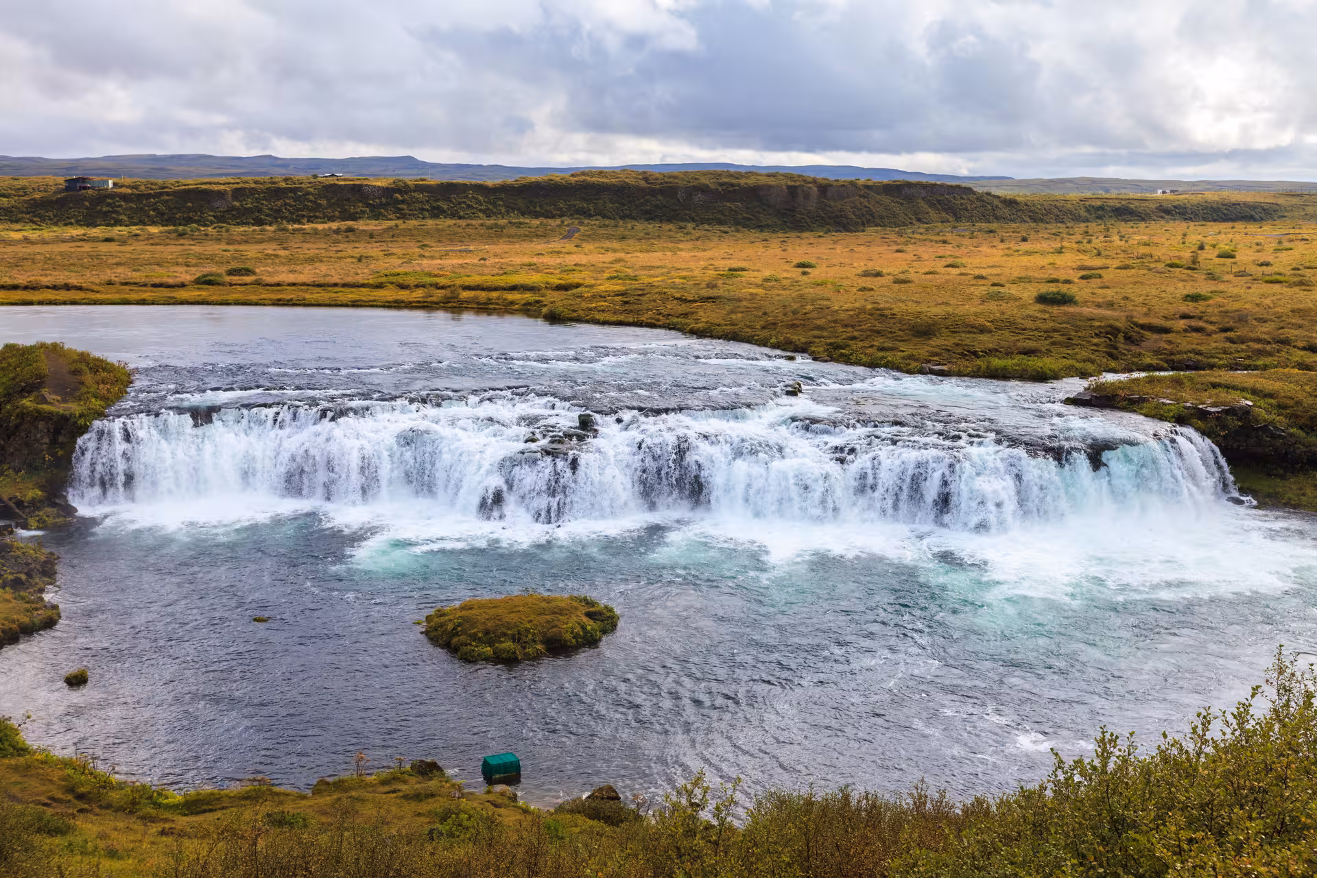 Wide cascading waterfall over volcanic rock ledge in Iceland's golden circle landscape under cloudy skies