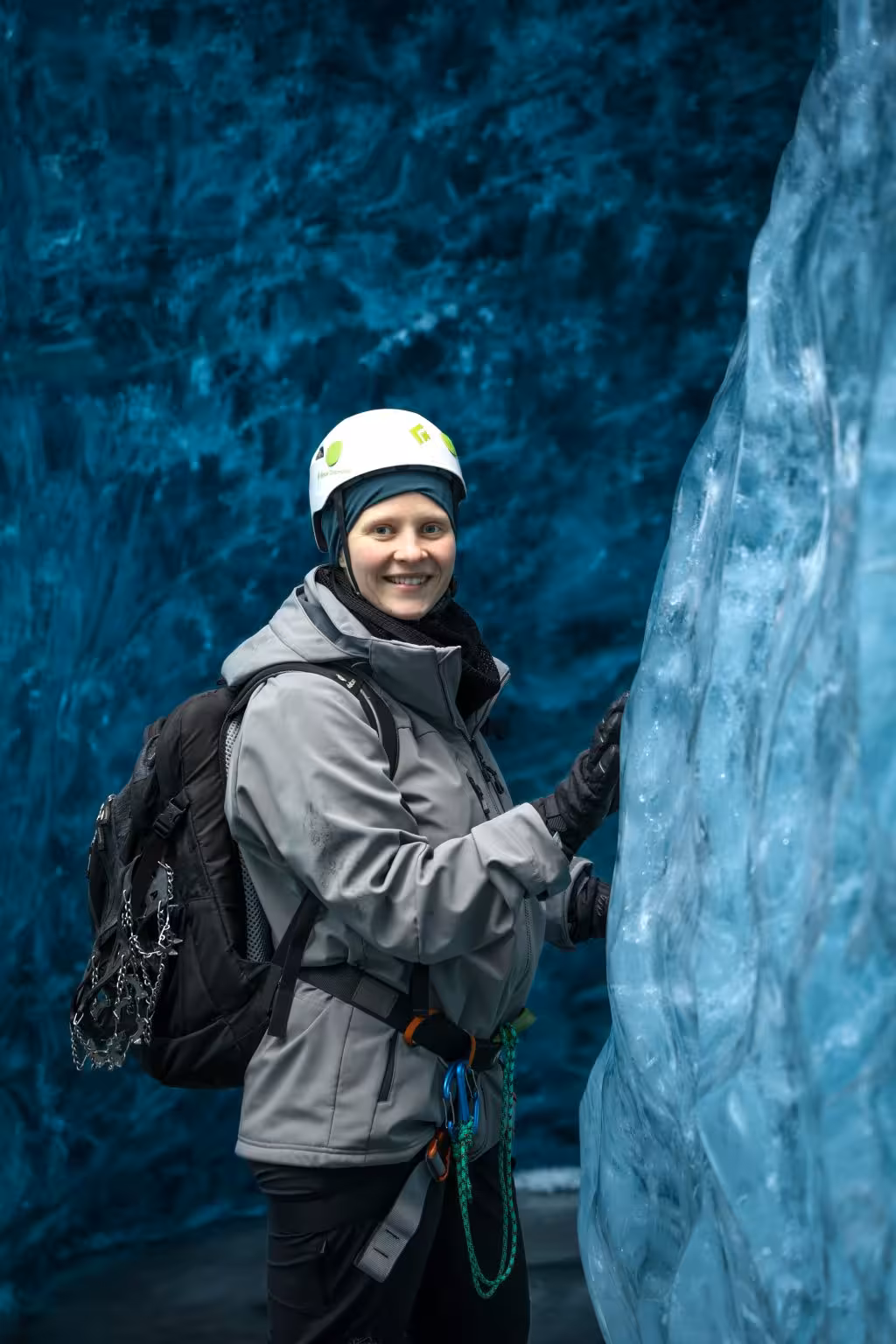 Smiling female hiker in helmet and crampons explores a vivid blue ice cave on Iceland glacier hike.