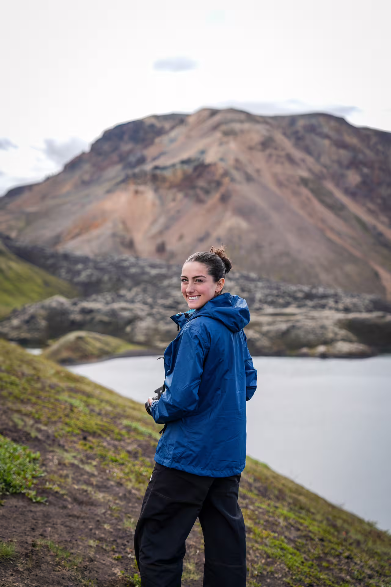 Smiling female hiker in blue jacket stands on a scenic highland trail beside a calm mountain lake in Iceland