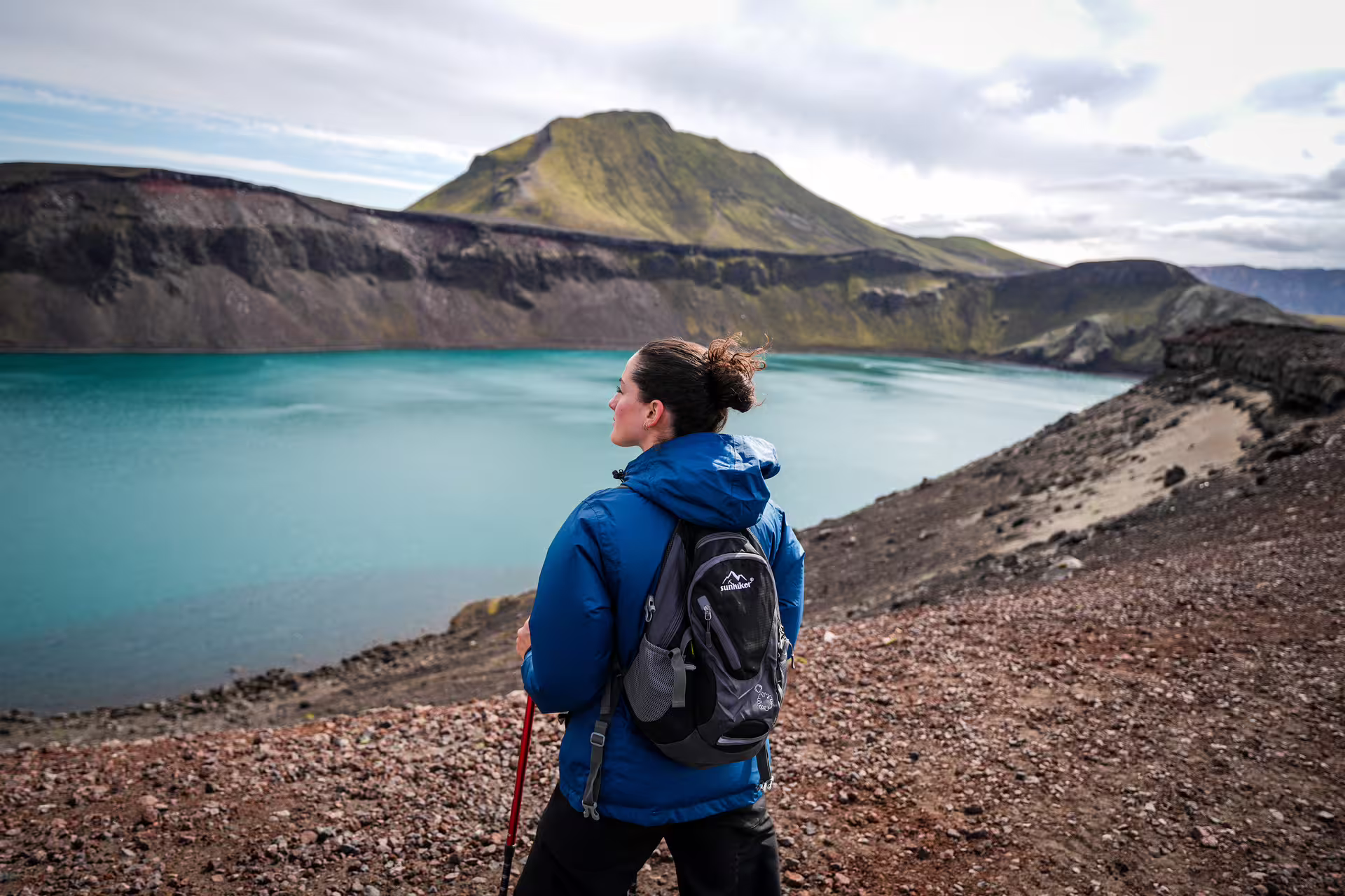 Female hiker with backpack and trekking pole gazes at a stunning turquoise crater lake on The Hidden Highlands Iceland tour