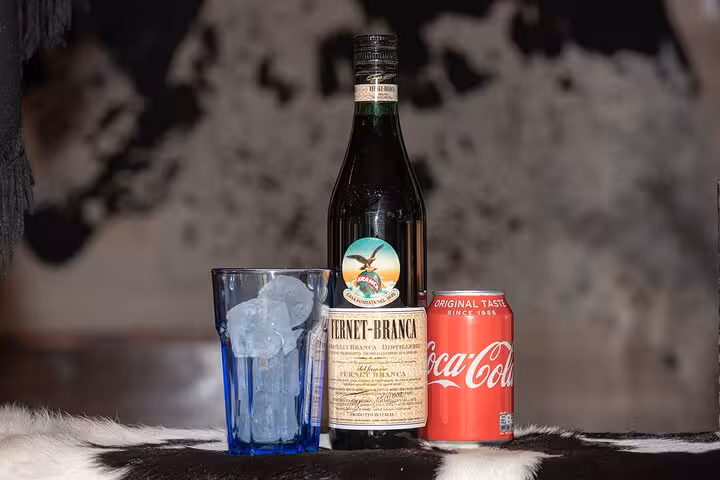 Fernet Branca bottle, a glass filled with ice cubes, and a can of Coca-Cola on a furry surface.