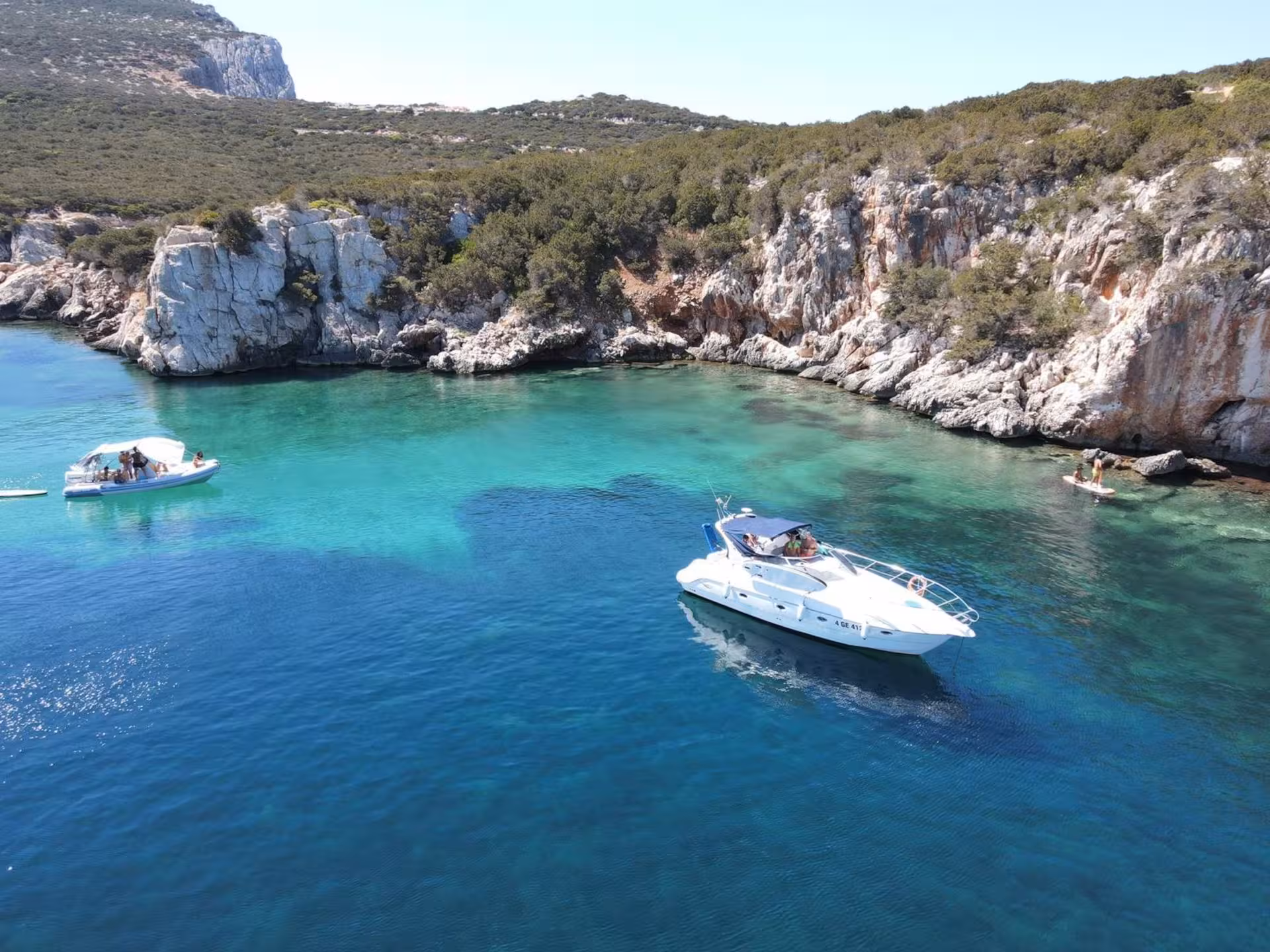 Boats anchored in the serene turquoise bay of the Gulf of Alghero, with rocky cliffs and vibrant greenery in the background.