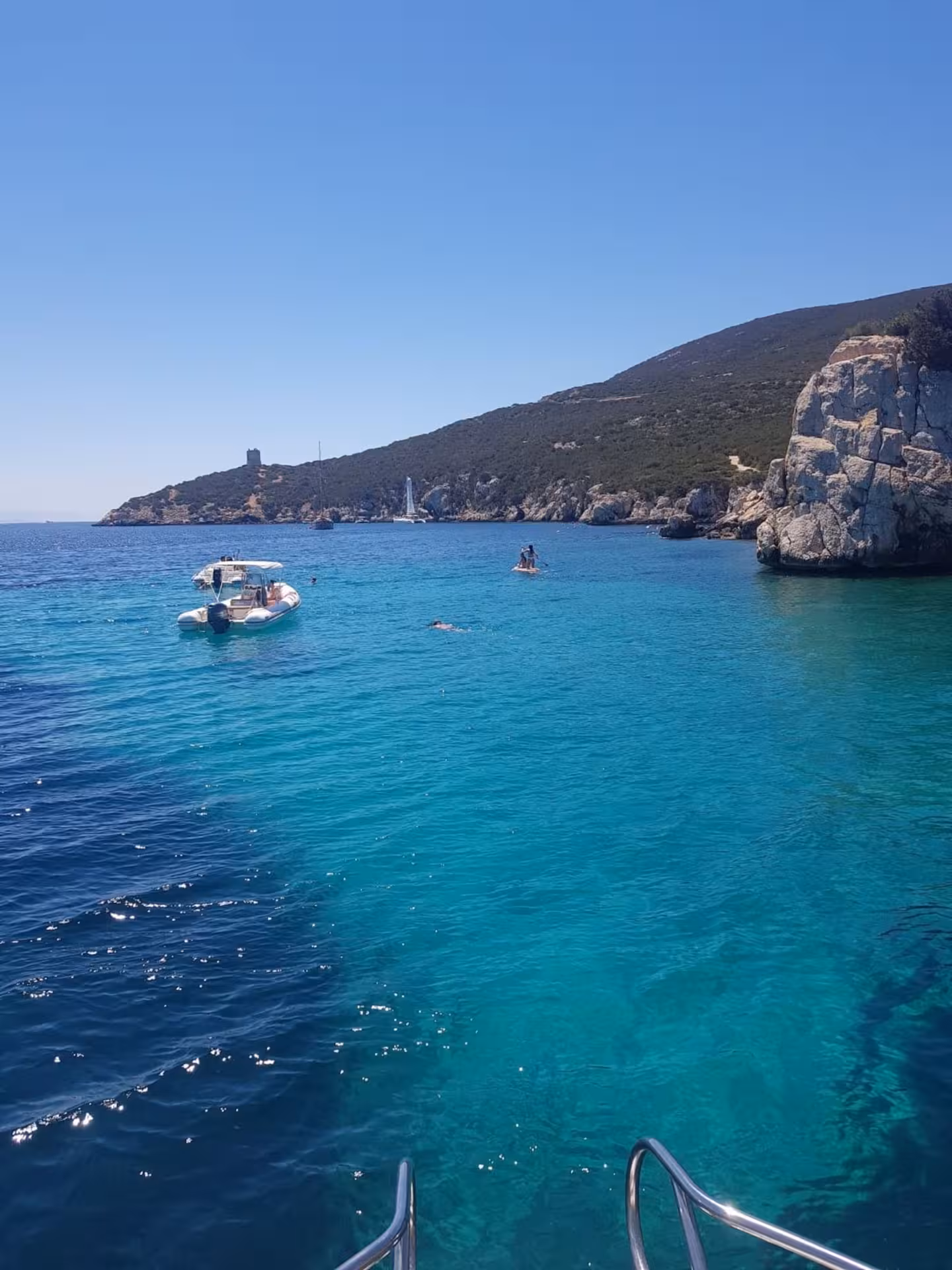 Scenic view of boats and swimmers in the turquoise waters of the Gulf of Alghero with rocky coastline.