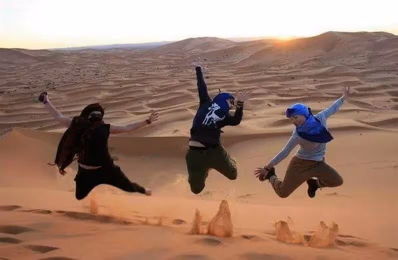 Three travelers joyfully jump on sand dunes at sunset in Merzouga Sahara during 4-day desert tour from Fes or Marrakech.