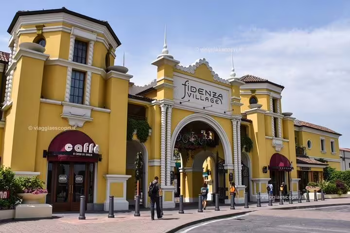 The grand entrance of Fidenza Village, a premier shopping destination near Milan, featuring vibrant yellow buildings and cafes.