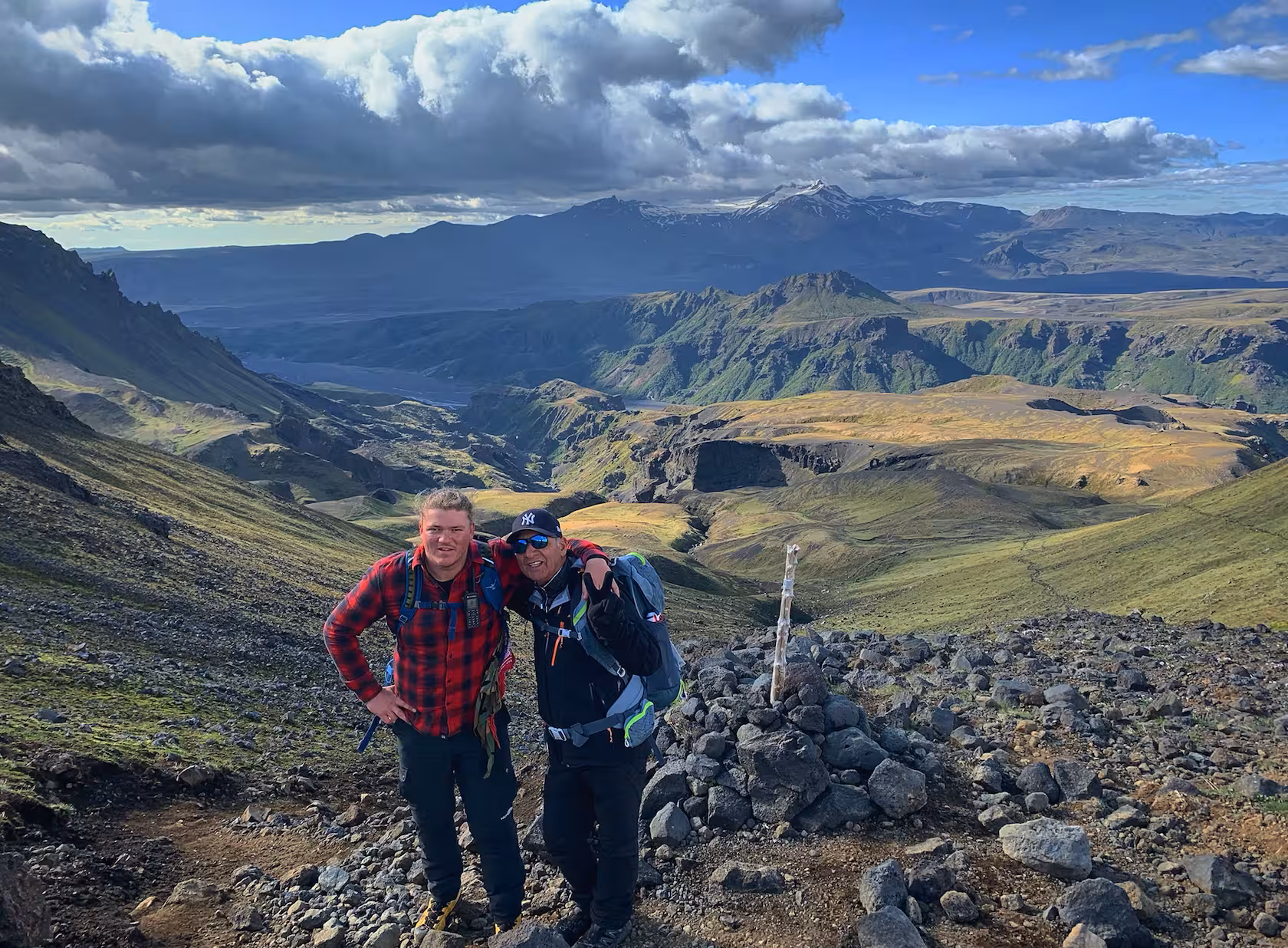 Two hikers posing at a rocky trail summit with sweeping green valley and snow-capped mountains in Iceland