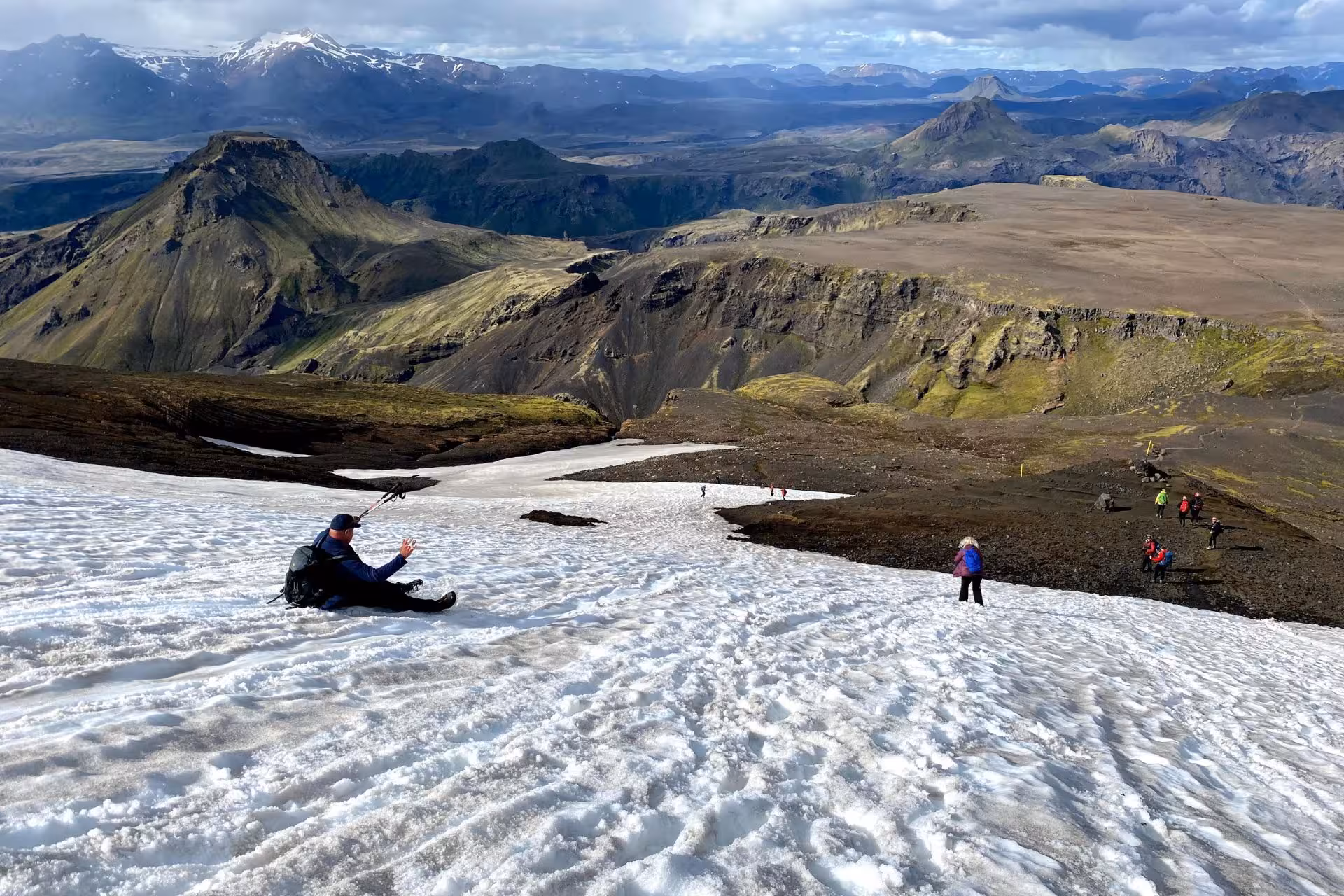 Hikers sliding and walking down a snowy slope on Fimmvörðuháls trail with Iceland's volcanic landscape behind