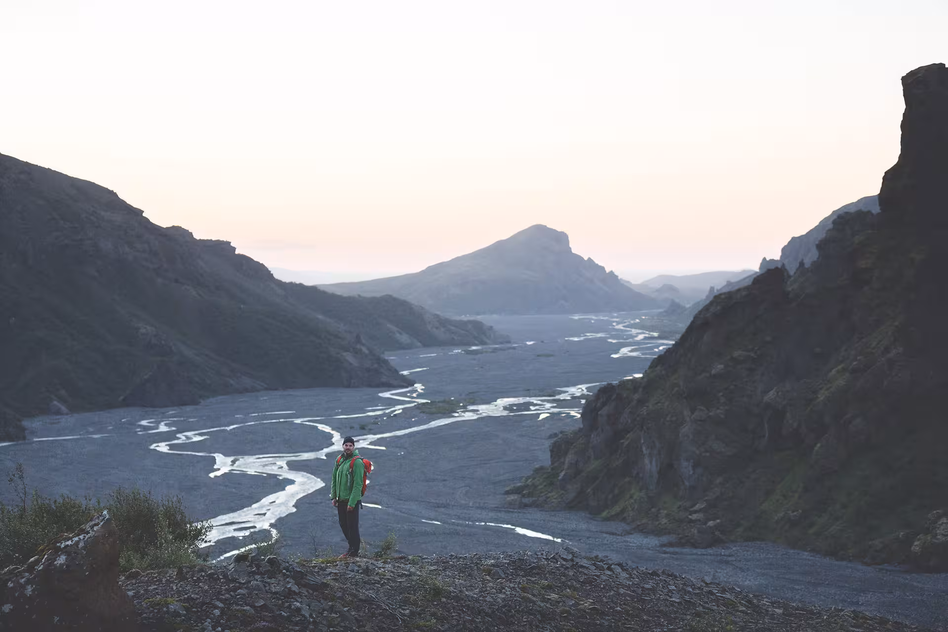 Hiker in green jacket standing on rocky ridge overlooking braided glacial river valley in Iceland at dusk