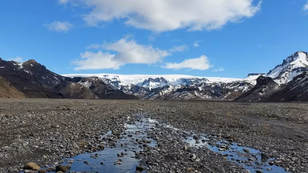 Rocky glacial valley with meltwater stream leading to snow-capped mountains under blue sky in Iceland