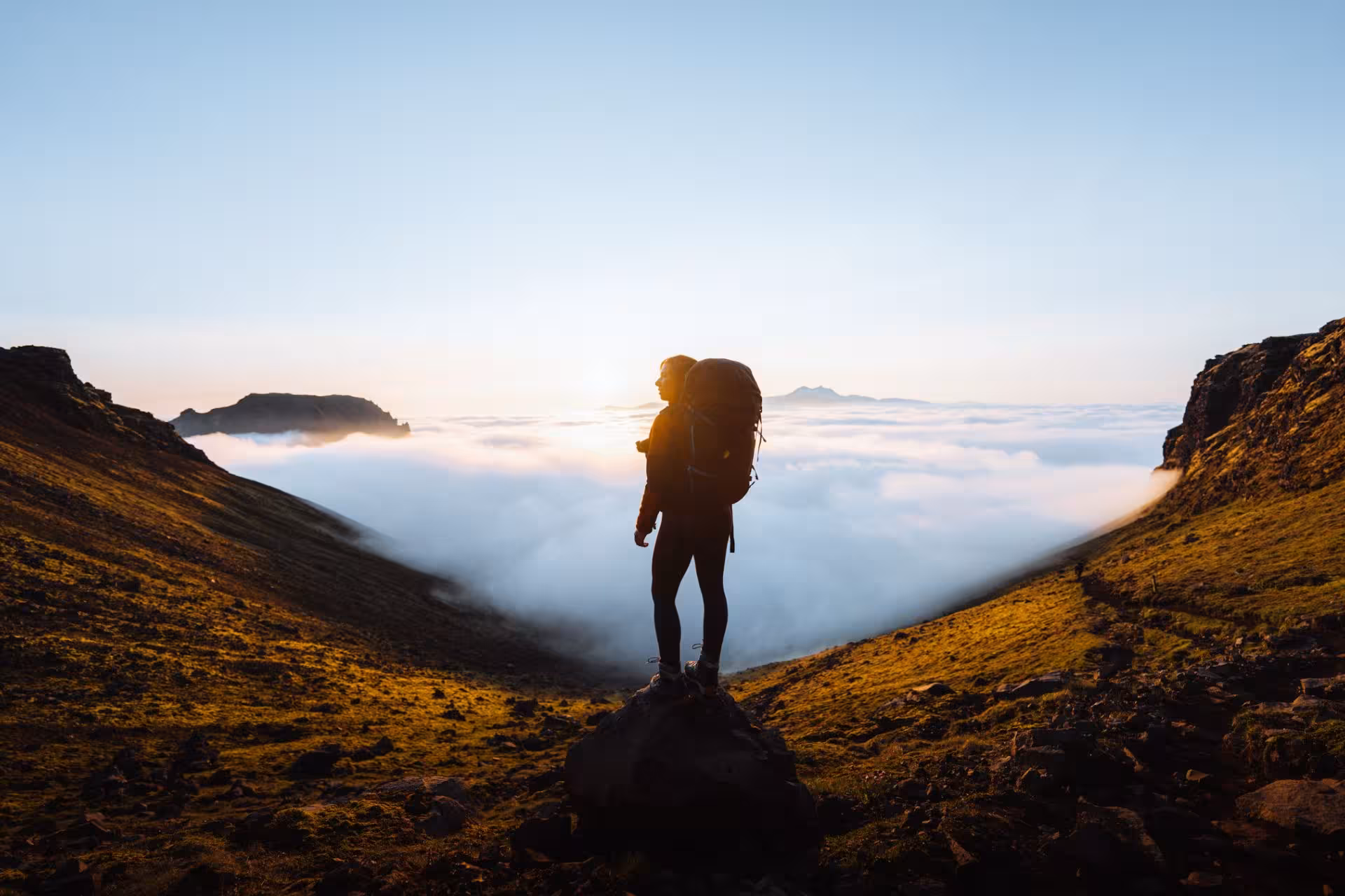 Hiker with backpack standing on rock above clouds at sunrise on Fimmvorduhals trail, Iceland