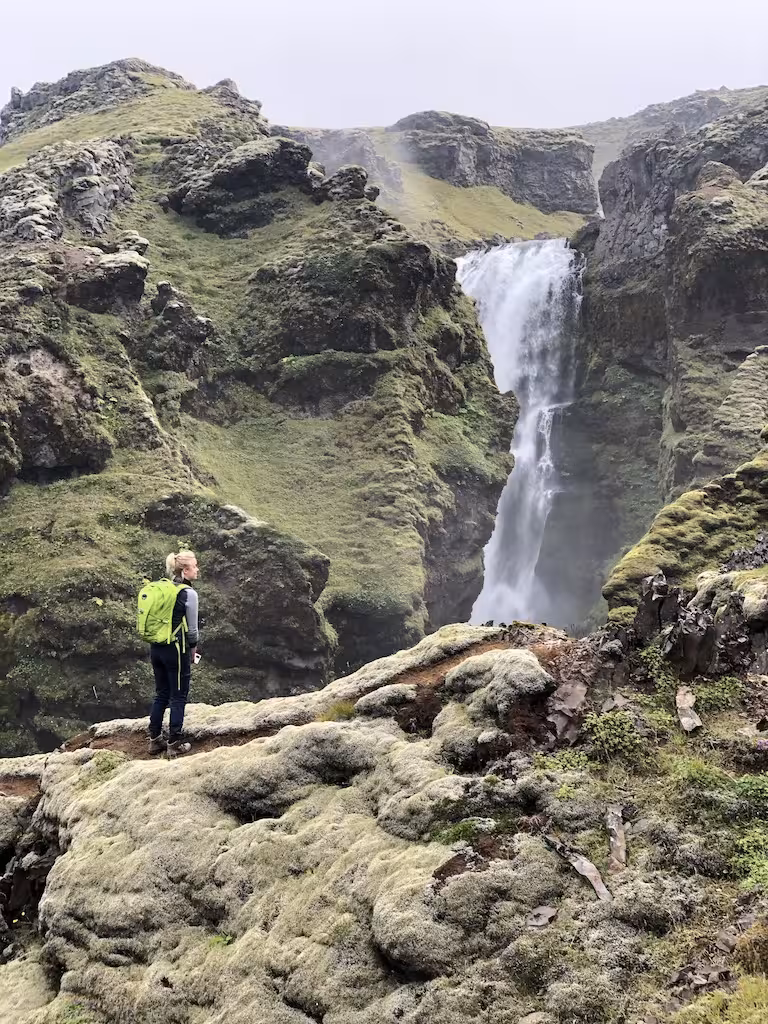 Hiker with green backpack standing on mossy rocks admiring waterfall on Fimmvorduhals trail Iceland