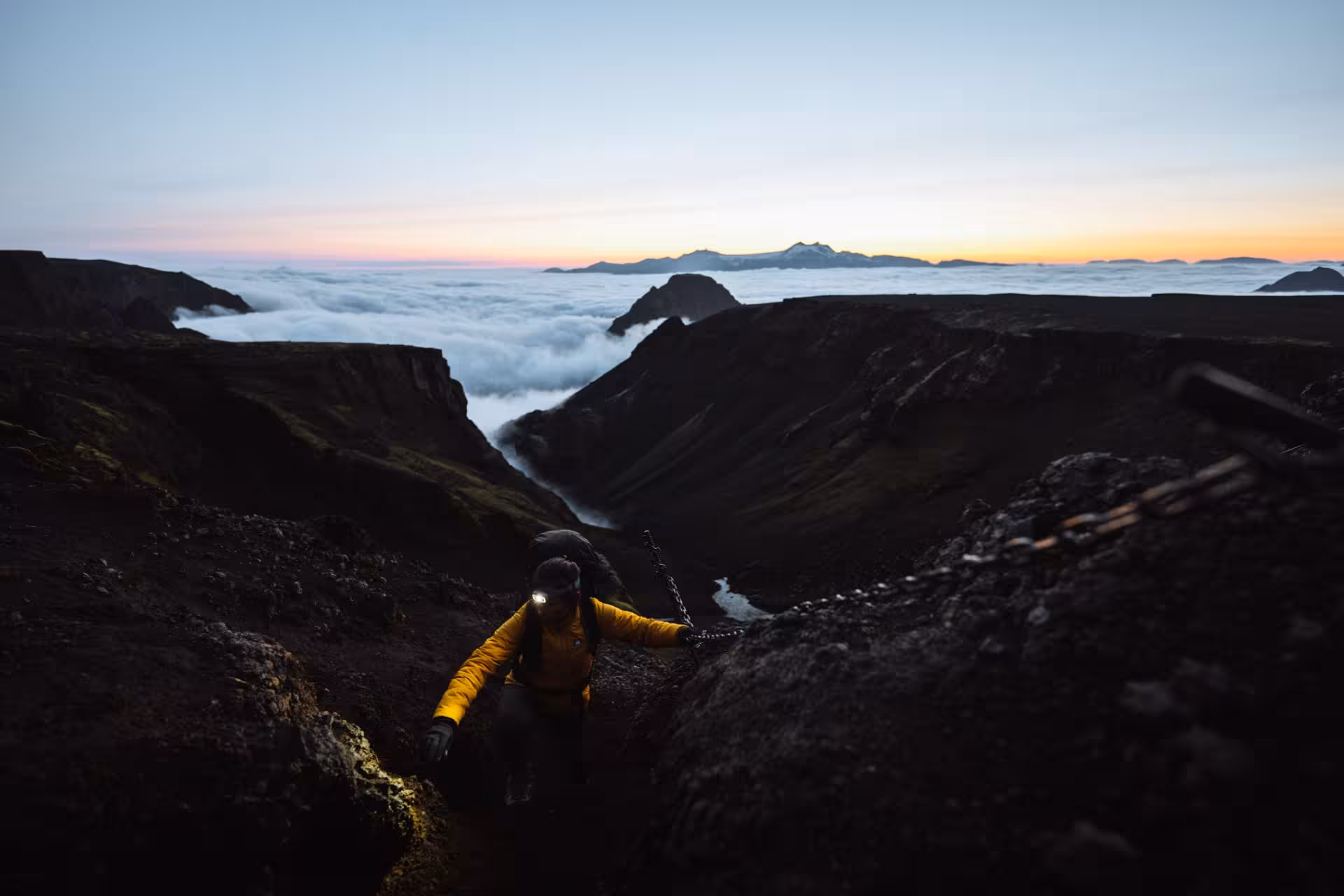 Hiker in yellow jacket climbing steep volcanic ridge at dawn above cloud sea on Fimmvorduhals trek Iceland