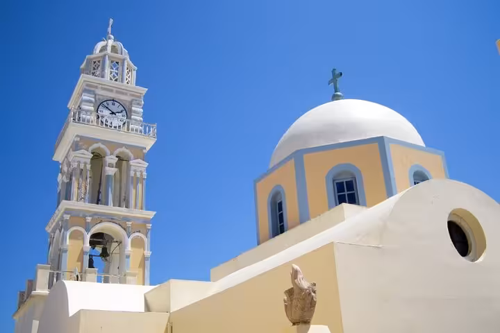 White domed Catholic church with ornate clock tower bell tower against blue sky in Fira, Santorini