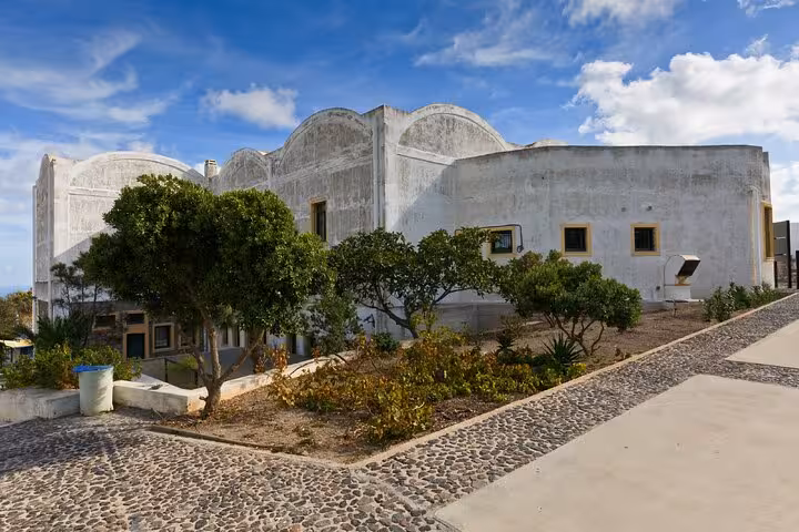 White-washed historic building with arched roofline and lush green trees in Fira, Santorini