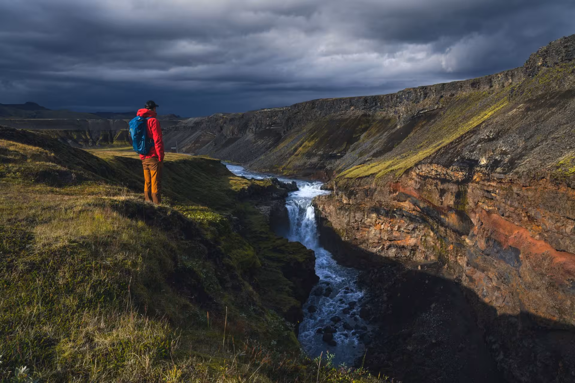 Hiker in red jacket overlooking a waterfall canyon in Fjallabak Nature Reserve under dramatic storm clouds