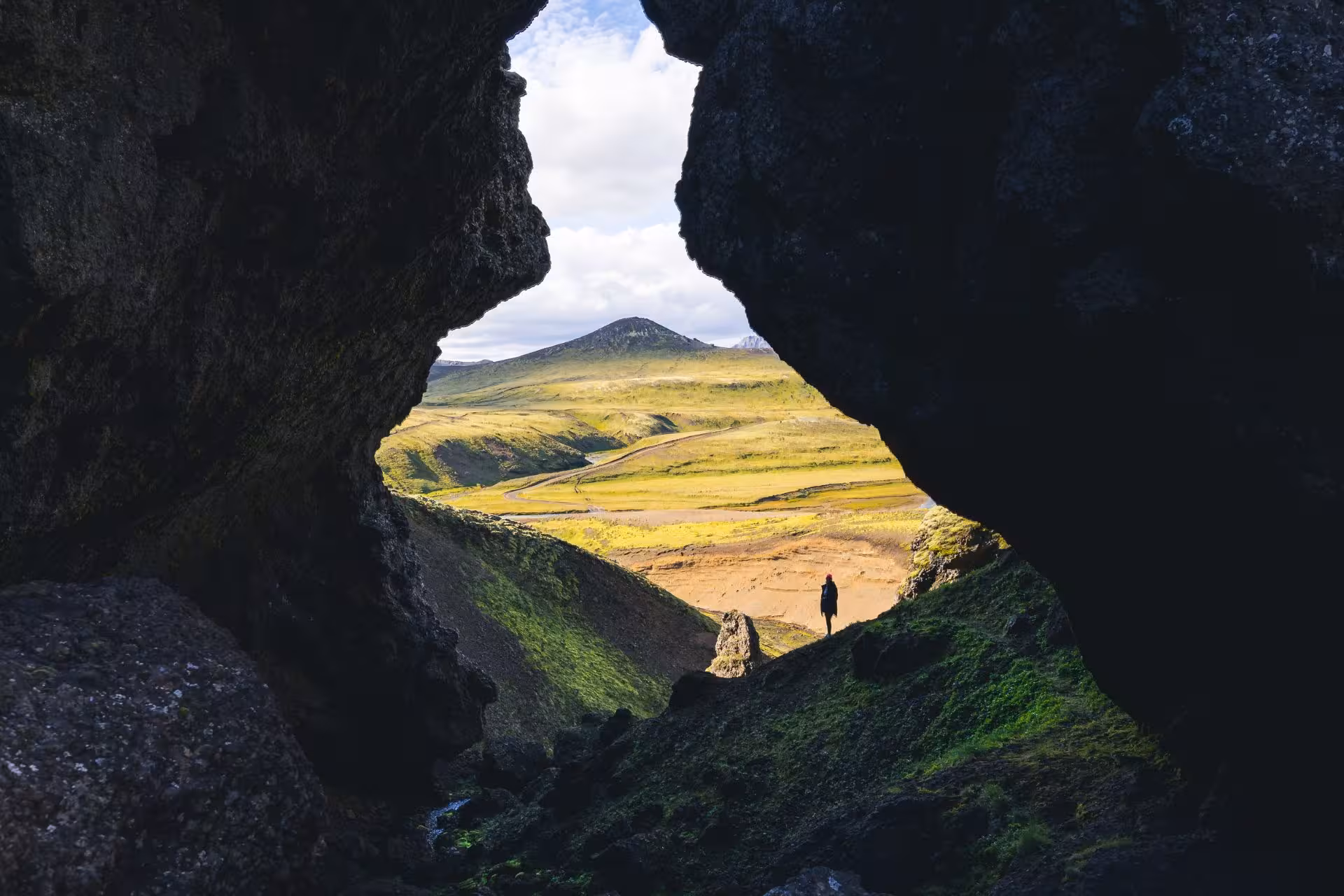 Silhouette of person standing at cave opening overlooking green Fjallabak landscape with volcanic mountain