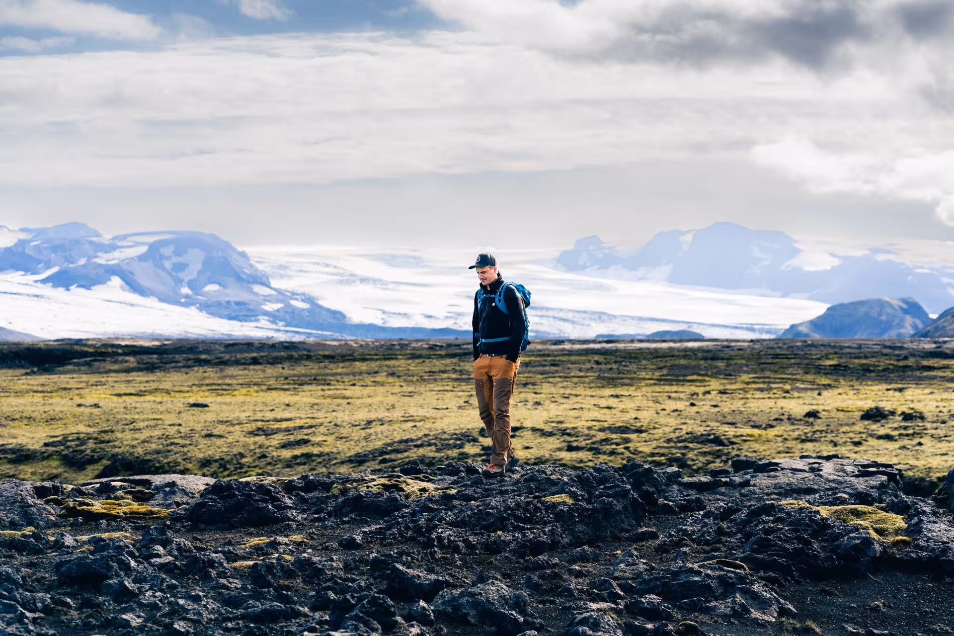 Hiker with backpack standing on volcanic lava field with snow-capped mountains and glacier in background, Iceland