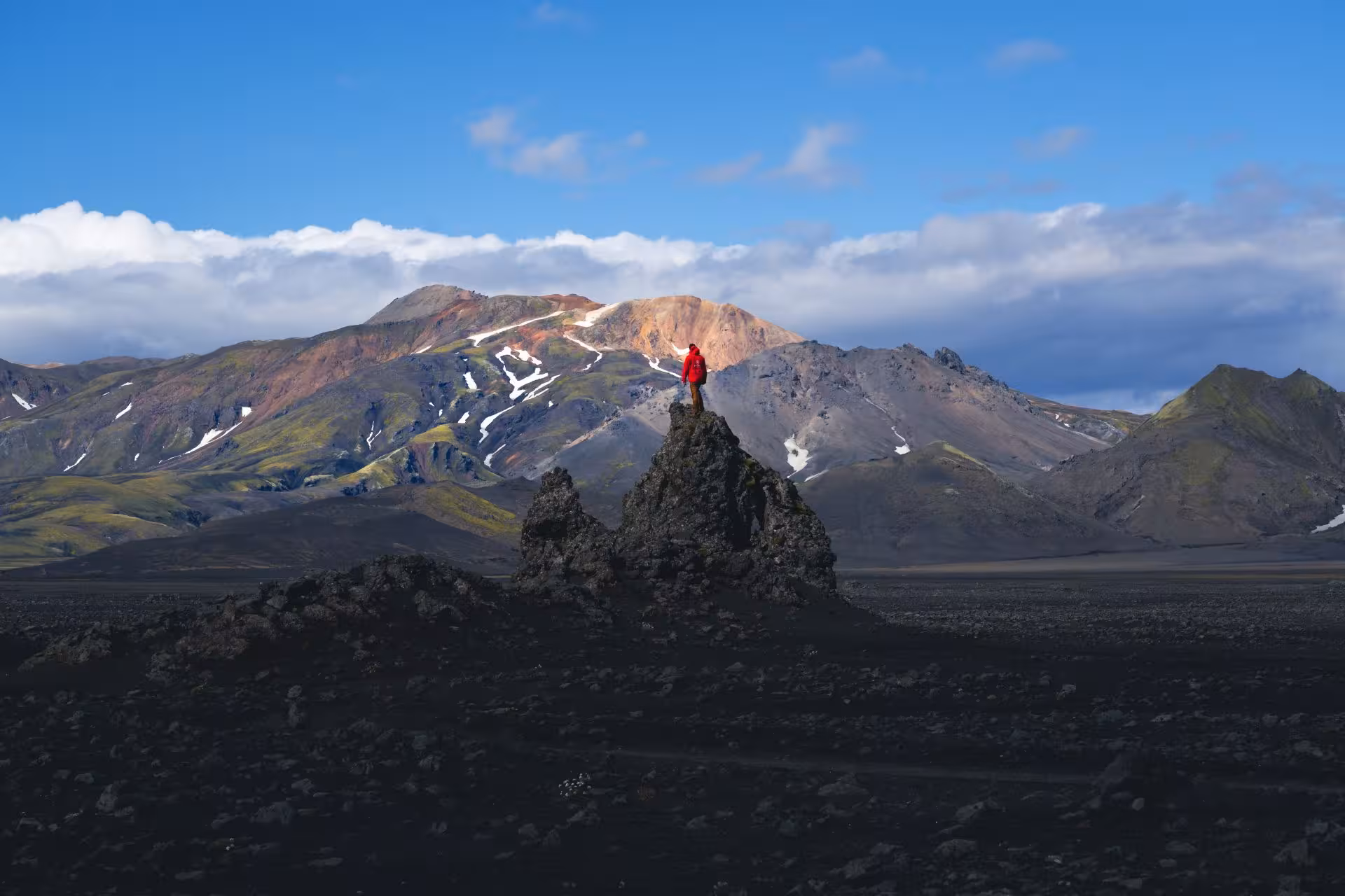 Hiker in red jacket standing on volcanic rock pinnacle with colorful Fjallabak mountains behind