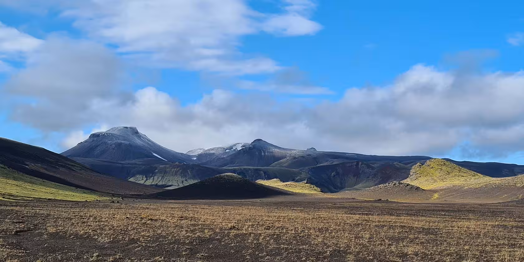 Vast volcanic plain with dark lava hills and snow-capped mountains under blue sky in Fjallabak Nature Reserve, Iceland