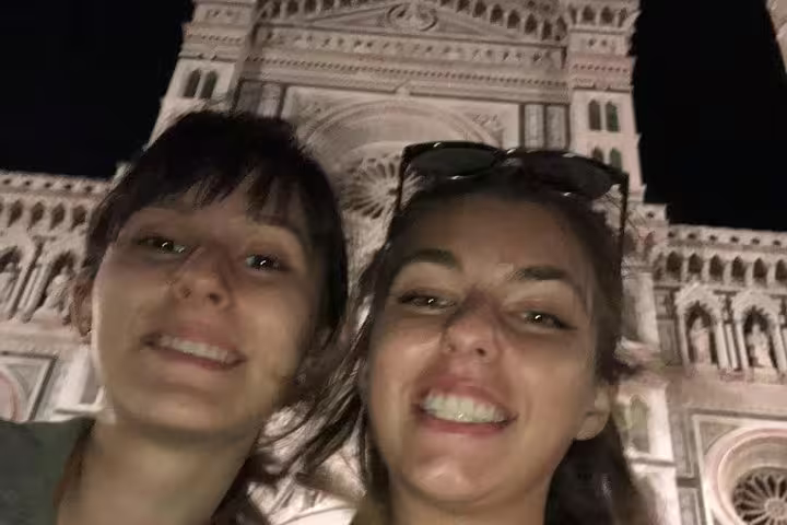 Two smiling women taking a selfie at night in front of the illuminated Florence Cathedral facade