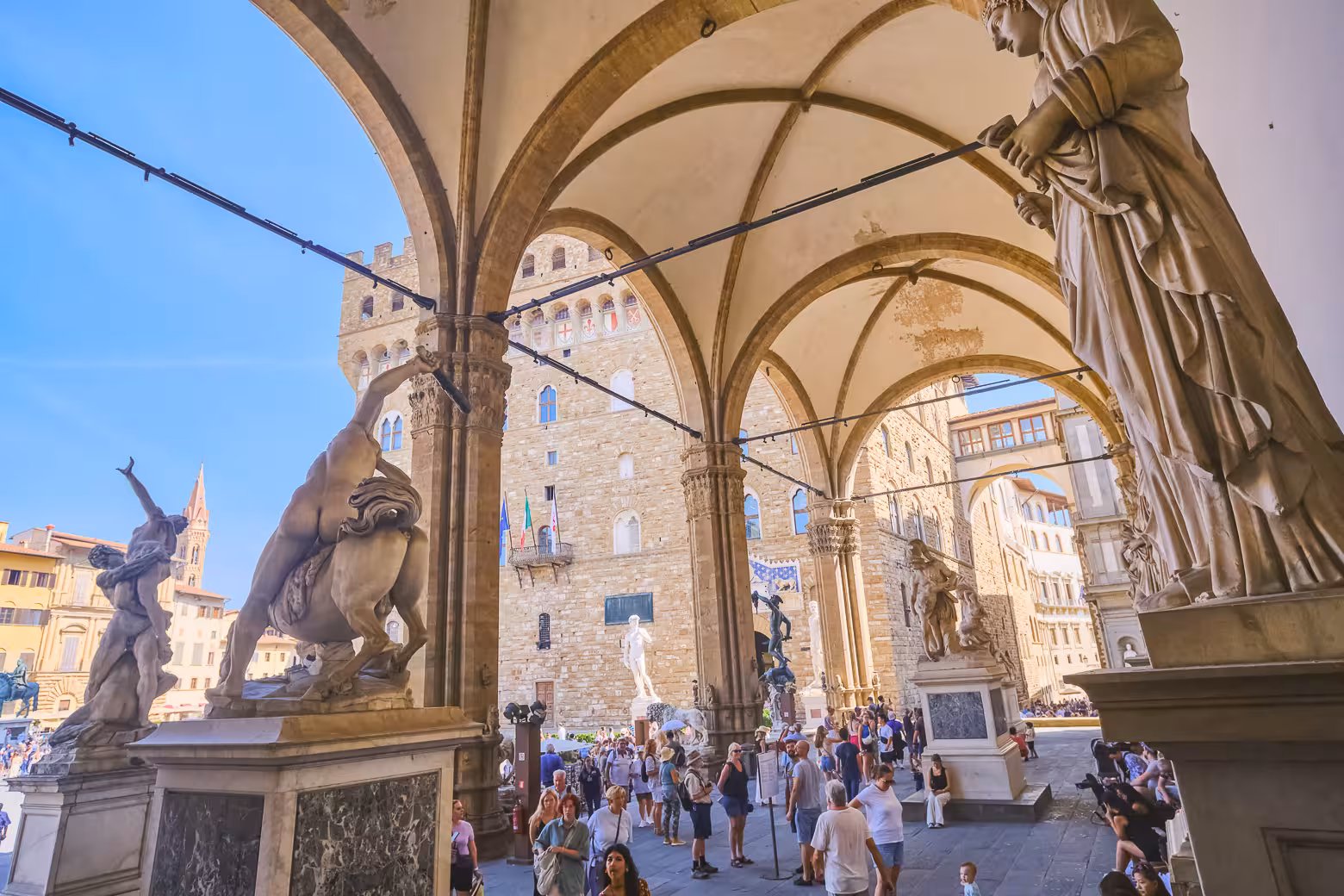 Tourists explore Florence's Piazza della Signoria, surrounded by historic statues and architecture on a sunny day.