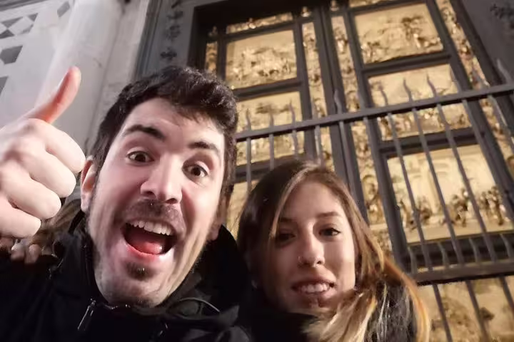 Two tourists smiling in front of Florence Baptistery golden Gates of Paradise at night