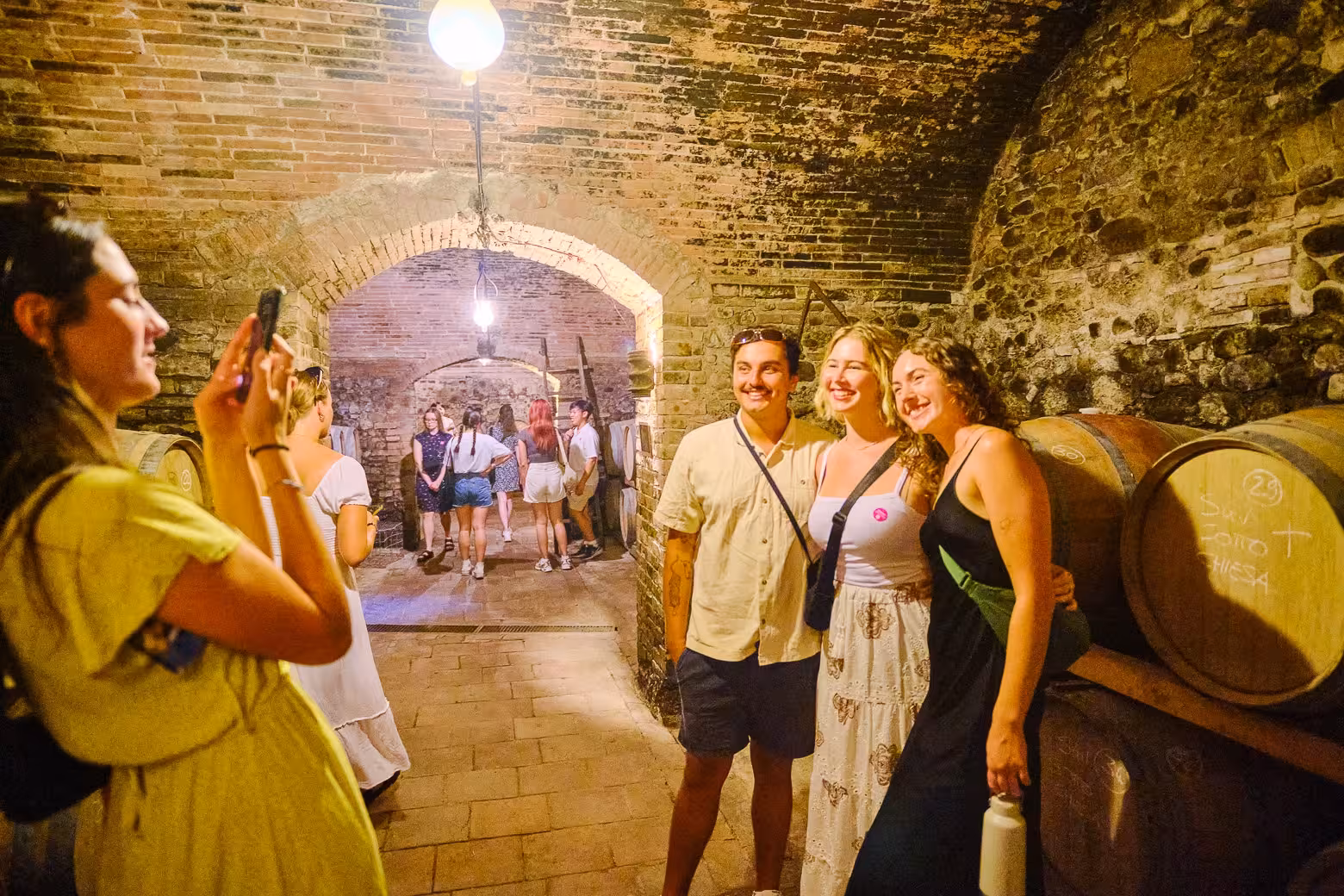 Visitors posing for photos during a wine tasting tour in a historic Florence cellar with large wine barrels.