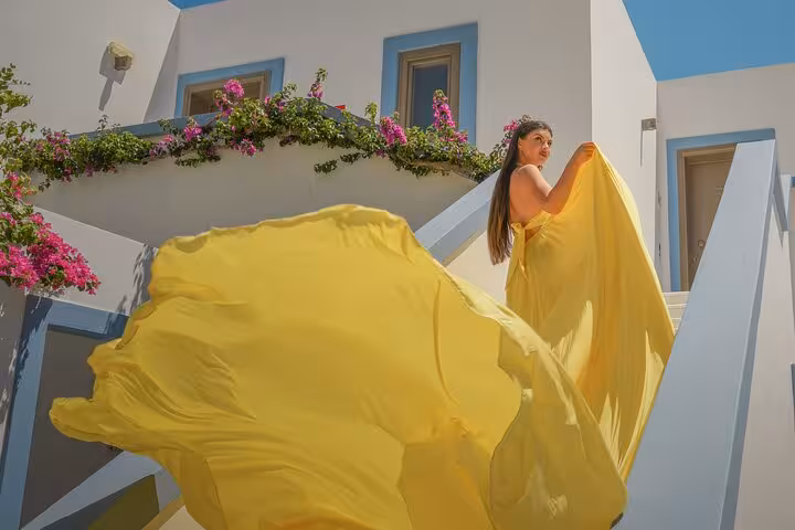 Woman in a flowing yellow dress poses on sunlit steps with blooming flowers in Hersonissos, ideal for a unique photoshoot.