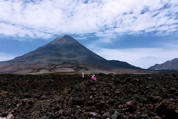 Hikers exploring the rocky terrain of Fogo Island with the towering volcano in the background under a vast sky.