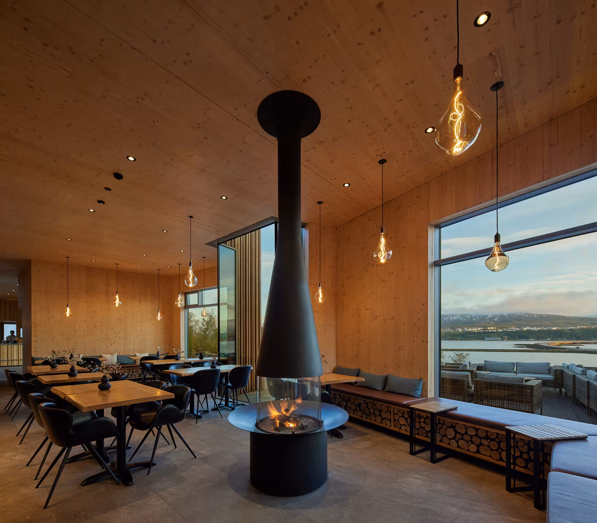 Warm wood-paneled dining room with central hanging fireplace and panoramic lake views at Forest Lagoon Akureyri