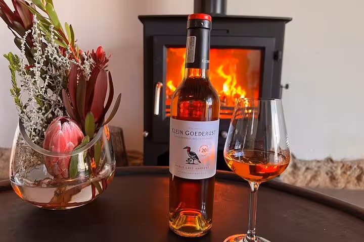 Cozy setting with Klein Goederust wine bottle and glass by a warm fireplace, perfect for a Franschhoek tour.