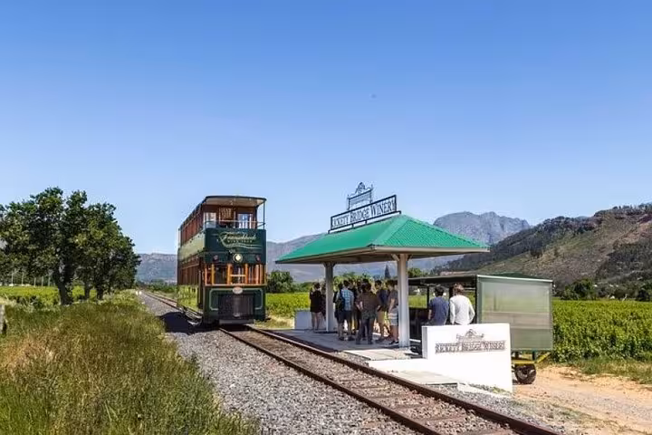 Franschhoek Wine Tram arriving at a picturesque vineyard station, surrounded by lush green fields and mountains.